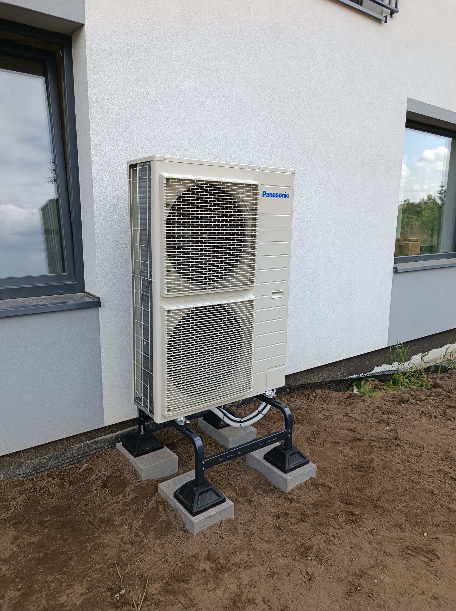 Zewnętrzna jednostka klimatyzacji Panasonic zamontowana na metalowej konstrukcji, posadowionej na betonowych bloczkach przy ścianie budynku z widocznymi oknami.