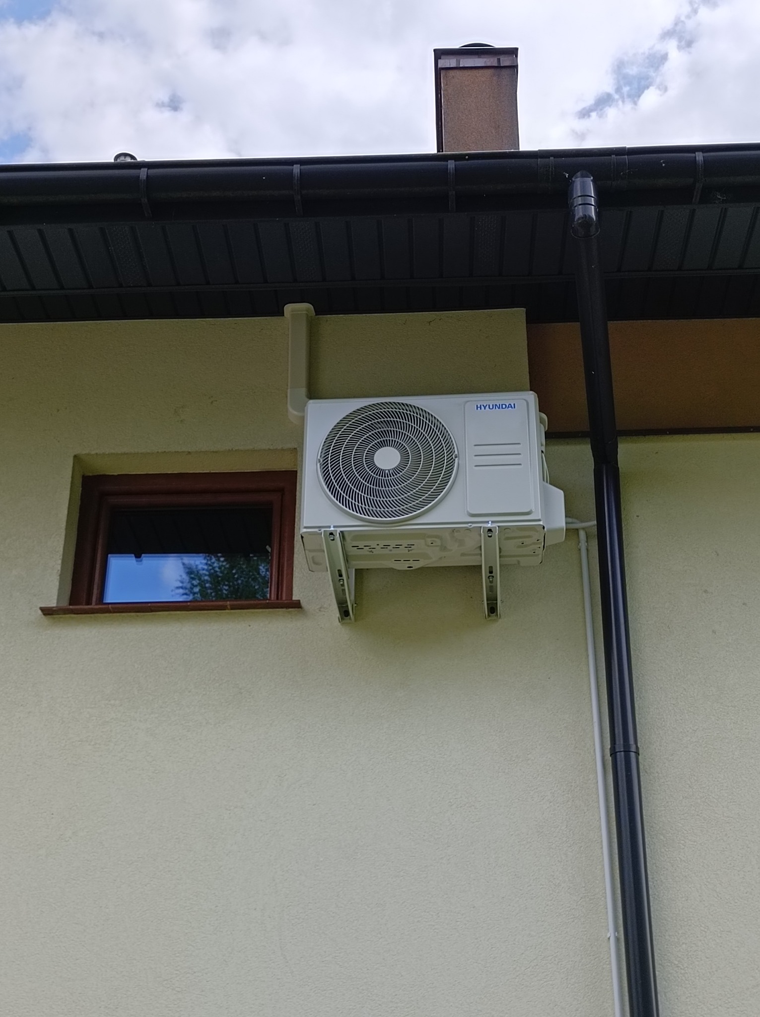 Zewnętrzna jednostka klimatyzacji Hyundai zamontowana na elewacji domu, widoczne rury odprowadzające skropliny i okno z brązową ramą.