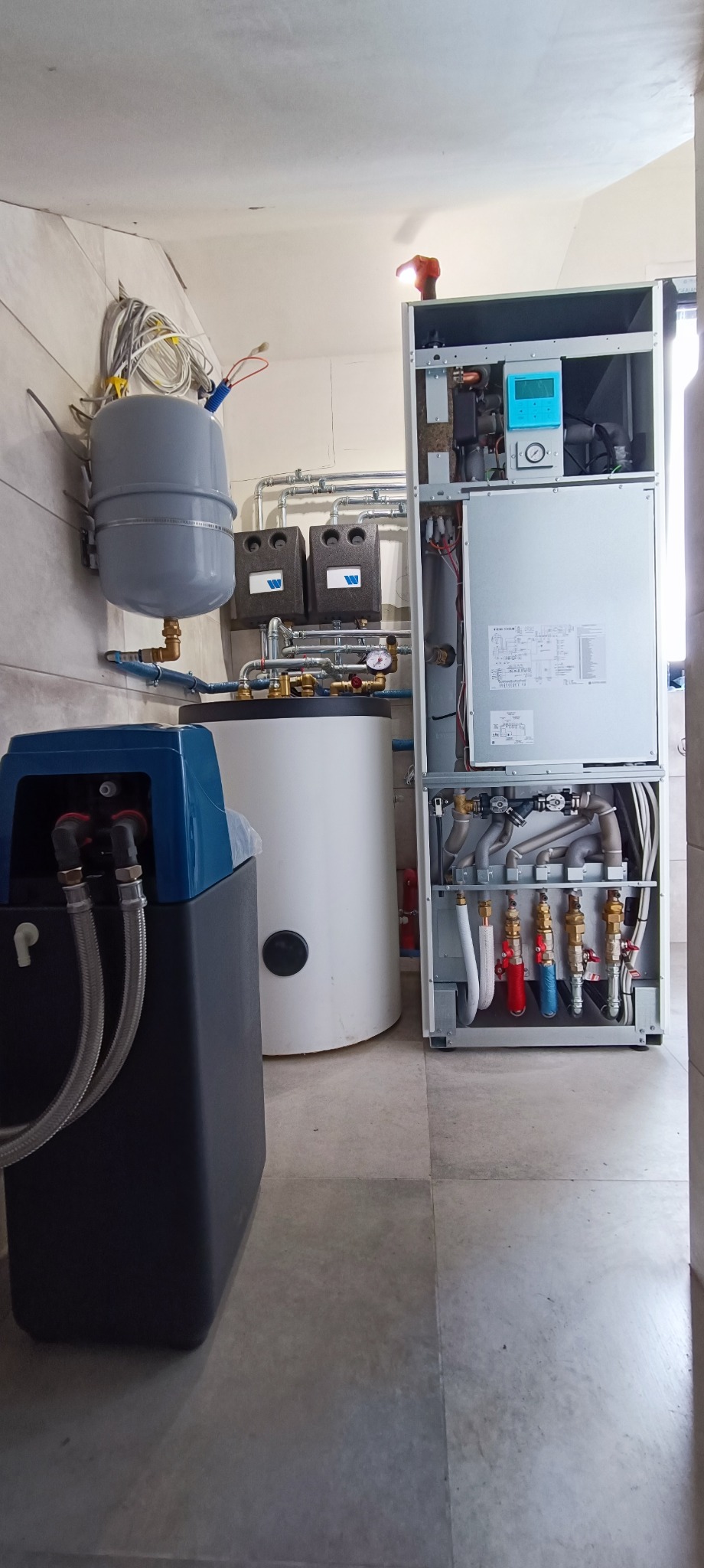 Kompletna instalacja grzewcza w kotłowni z widocznym zasobnikiem ciepłej wody, zbiornikiem wyrównawczym, zmiękczaczem wody oraz rozdzielaczem z zaworami, wszystko w pomieszczeniu wyłożonym płytkami.