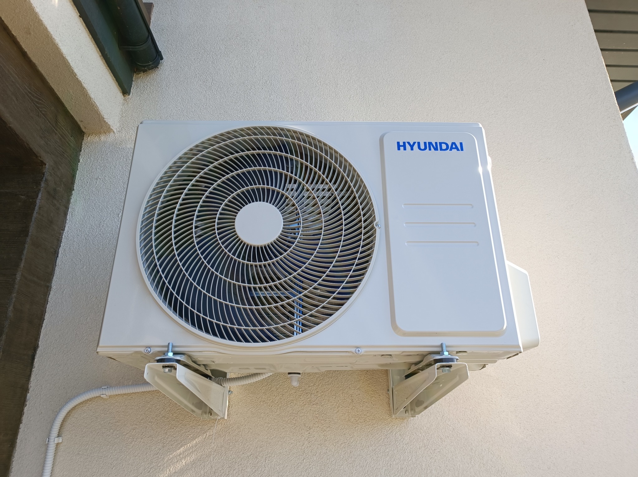 Biała jednostka zewnętrzna klimatyzatora marki Hyundai zamontowana na jasnej, teksturowanej ścianie budynku z widocznymi wspornikami montażowymi i rurami.