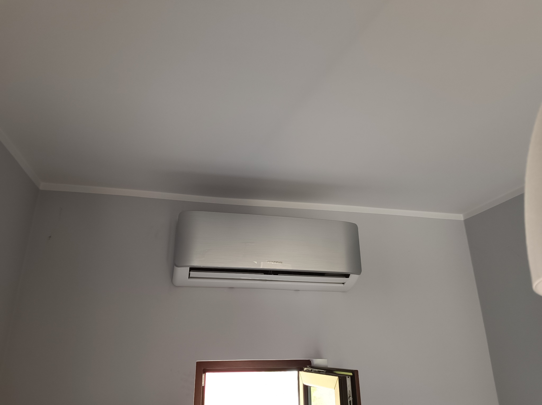 Zamontowany na szarej ścianie, srebrny klimatyzator marki Hyundai nad otwartym oknem w ramie z ciemnego drewna.