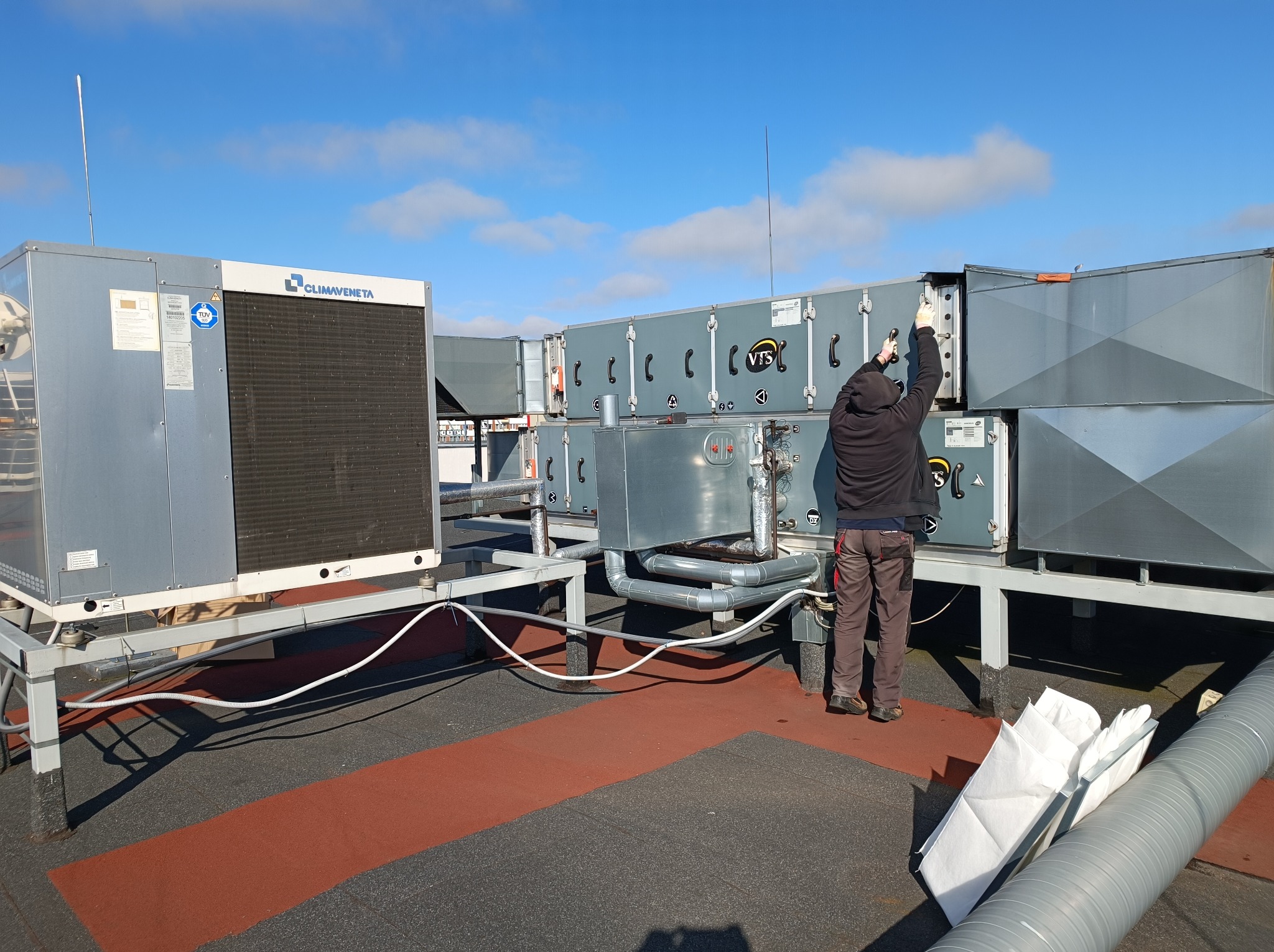 Instalator w rękawicach montuje system wentylacyjny VTS na dachu budynku, widoczny również agregat klimatyzacyjny Climaveneta i fragment instalacji rurowej.