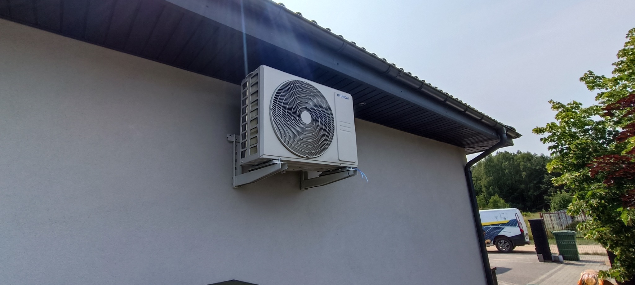 Zewnętrzna jednostka klimatyzacji Hyundai zamontowana na elewacji domu z widocznymi przewodami i fragmentem dachu z rynną, w tle częściowo widoczny samochód serwisowy.