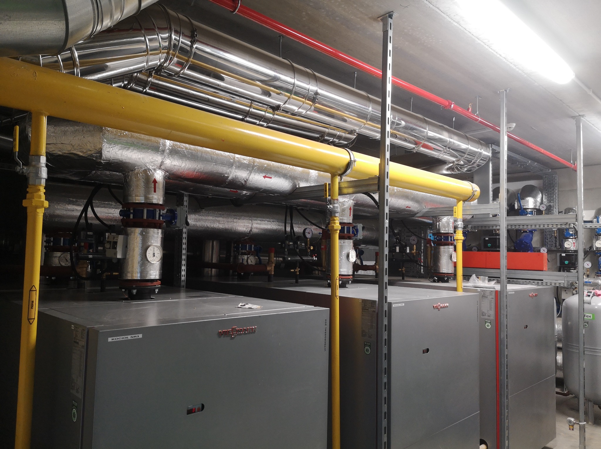 Kotłownia przemysłowa z czterema kotłami Viessmann Vitocrossal 200, rurami w kolorze żółtym, srebrnym i czerwonym pod sufitem, manometry na instalacji.
