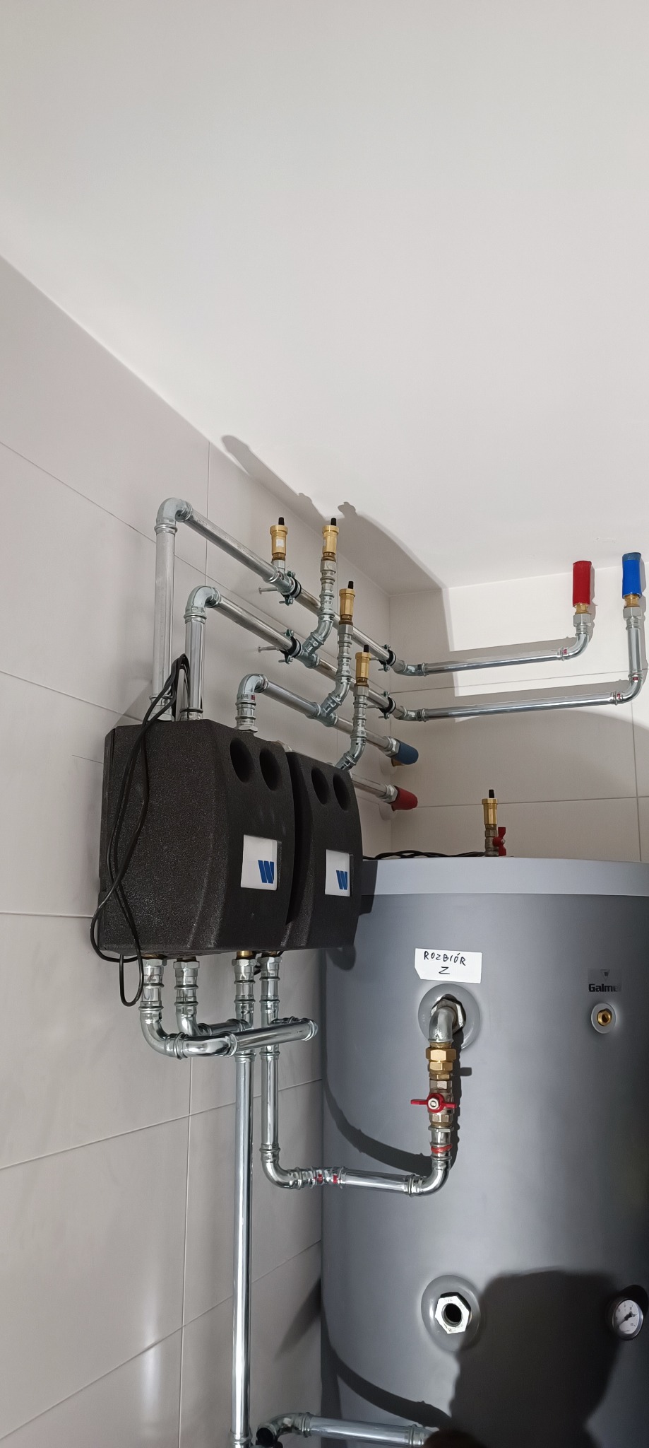 Instalacja grzewcza z szarym zbiornikiem i przewodami, widoczne zawory z czerwonymi i niebieskimi pokrętłami, oraz dwa czarne moduły z logo na ścianie z płytek.