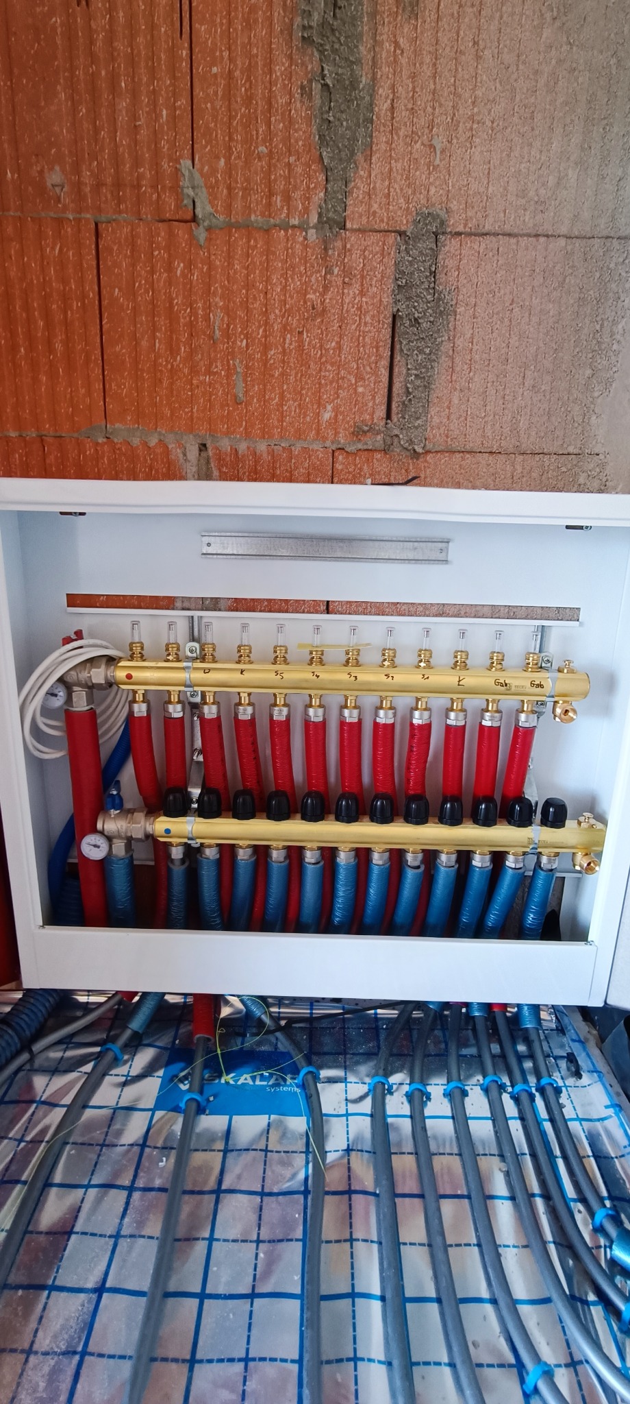 Rozdzielacz ogrzewania podłogowego w szafce instalacyjnej, widoczne rury czerwone i niebieskie, oraz szare rury ogrzewania podłogowego na folii z nadrukowaną siatką.