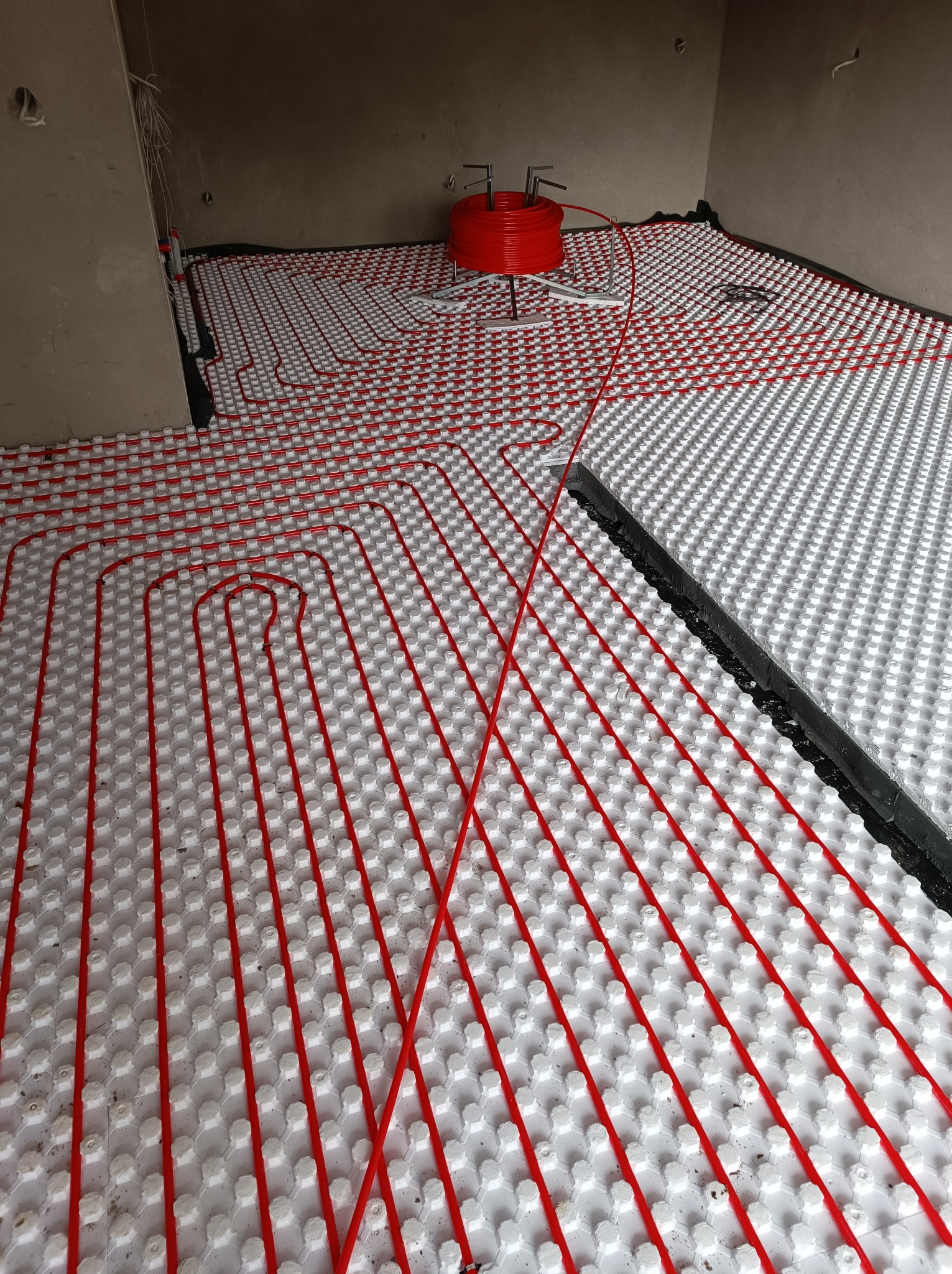 Instalacja ogrzewania podłogowego z czerwonymi rurkami na białych panelach izolacyjnych, widoczna rolka rur w tle, surowy stan wykończenia wnętrza.