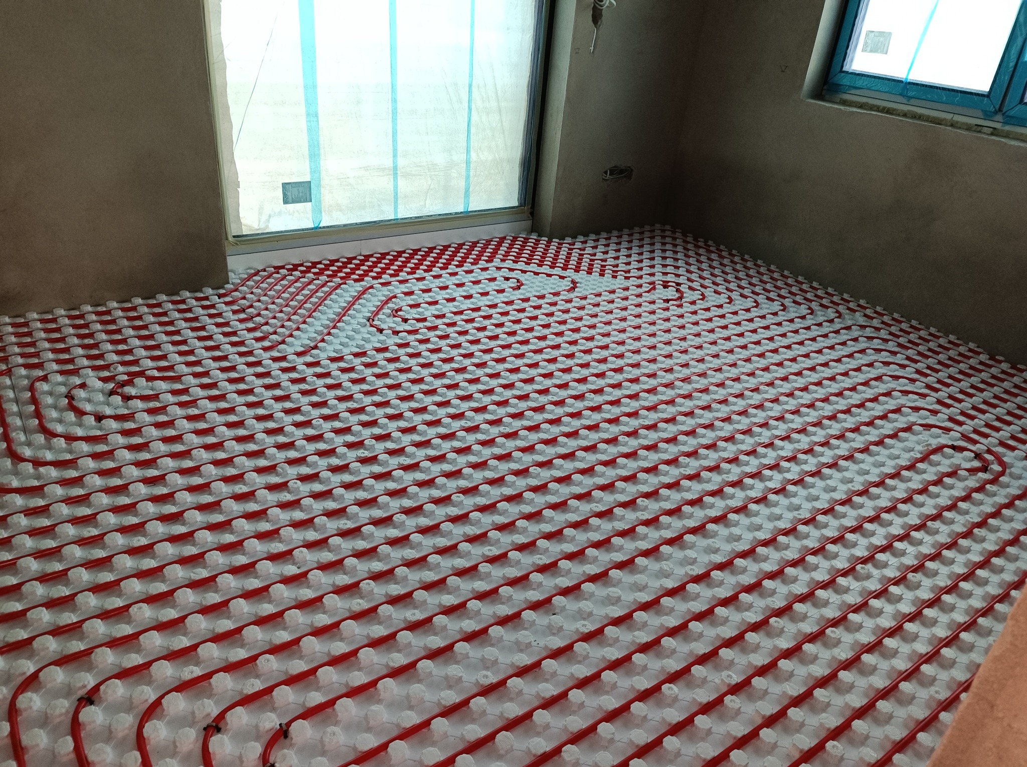 Czerwone rury ogrzewania podłogowego ułożone spiralnie na białych izolatorach, widok z góry na surową posadzkę w trakcie montażu.