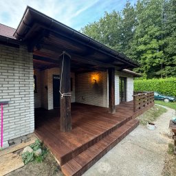 Jakub Kula - Zadaszony taras z drewnianą podłogą i schodami, wsparty na drewnianych kolumnach, z widoczną ścianą budynku z białej cegły i zielonym tłem drzew. Na pierwszym planie fragment betonowego podłoża.