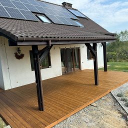 Jakub Kula - Nowoczesny dom z brązowym dachem i panelami słonecznymi, z drewnianym tarasem zadaszonym belkami, widok na ogród i wzgórza.