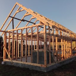 Jakub Kula - Konstrukcja szkieletowa budynku w trakcie budowy, widoczne drewniane elementy nośne i fundament z bloczków betonowych. W tle budynek i drzewa.