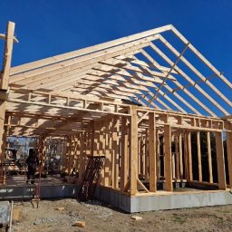 Jakub Kula - Surowa konstrukcja domu szkieletowego w trakcie budowy, widoczne drewniane ramy ścian i dachu na betonowej podstawie, błękitne niebo w tle, wczesny etap prac.