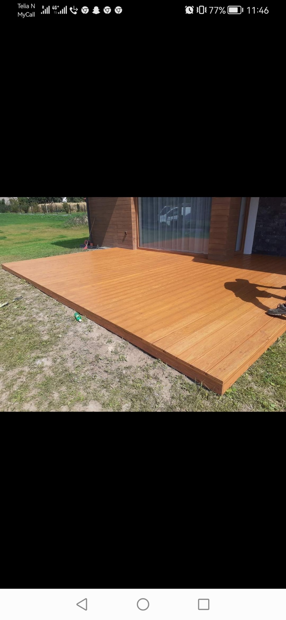 Nowy, duży taras z desek w odcieniach brązu, przylegający do budynku z dużym oknem, cień osoby w prawym górnym rogu, trawnik w tle.