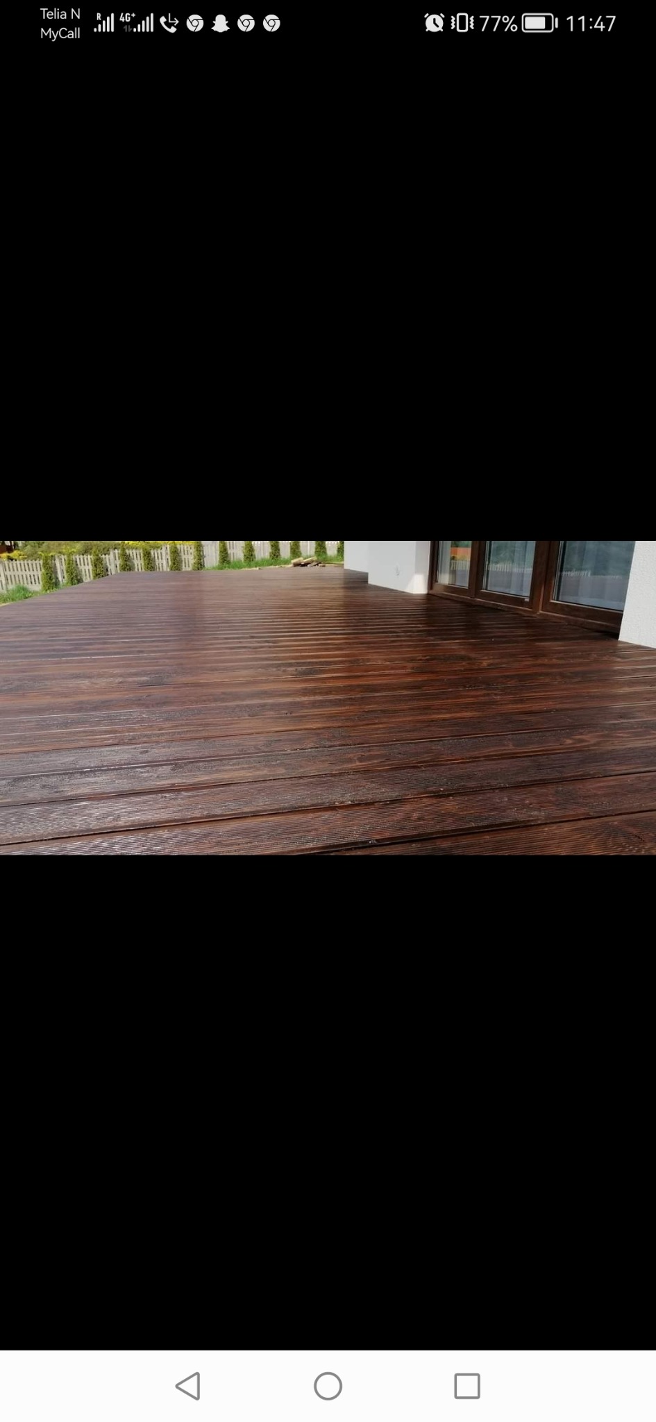 Ciemnobrązowy taras z desek, widok z poziomu podłogi, z przesuwnymi drzwiami balkonowymi i białym ogrodzeniem w tle.