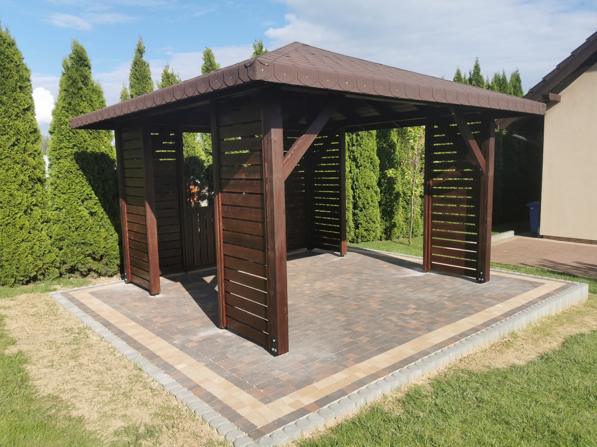 Ciemnobrązowa, drewniana pergola z zadaszeniem z gontu bitumicznego, otoczona tujami, z wybrukowaną podłogą z kostki w odcieniach brązu i beżu.