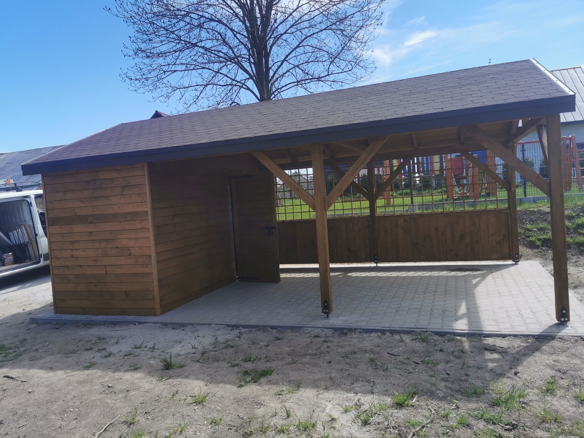 Drewniana konstrukcja z zadaszeniem z gontu bitumicznego, częściowo zabudowana, z otwartą przestrzenią i ażurowymi ścianami, posadowiona na kostce brukowej.