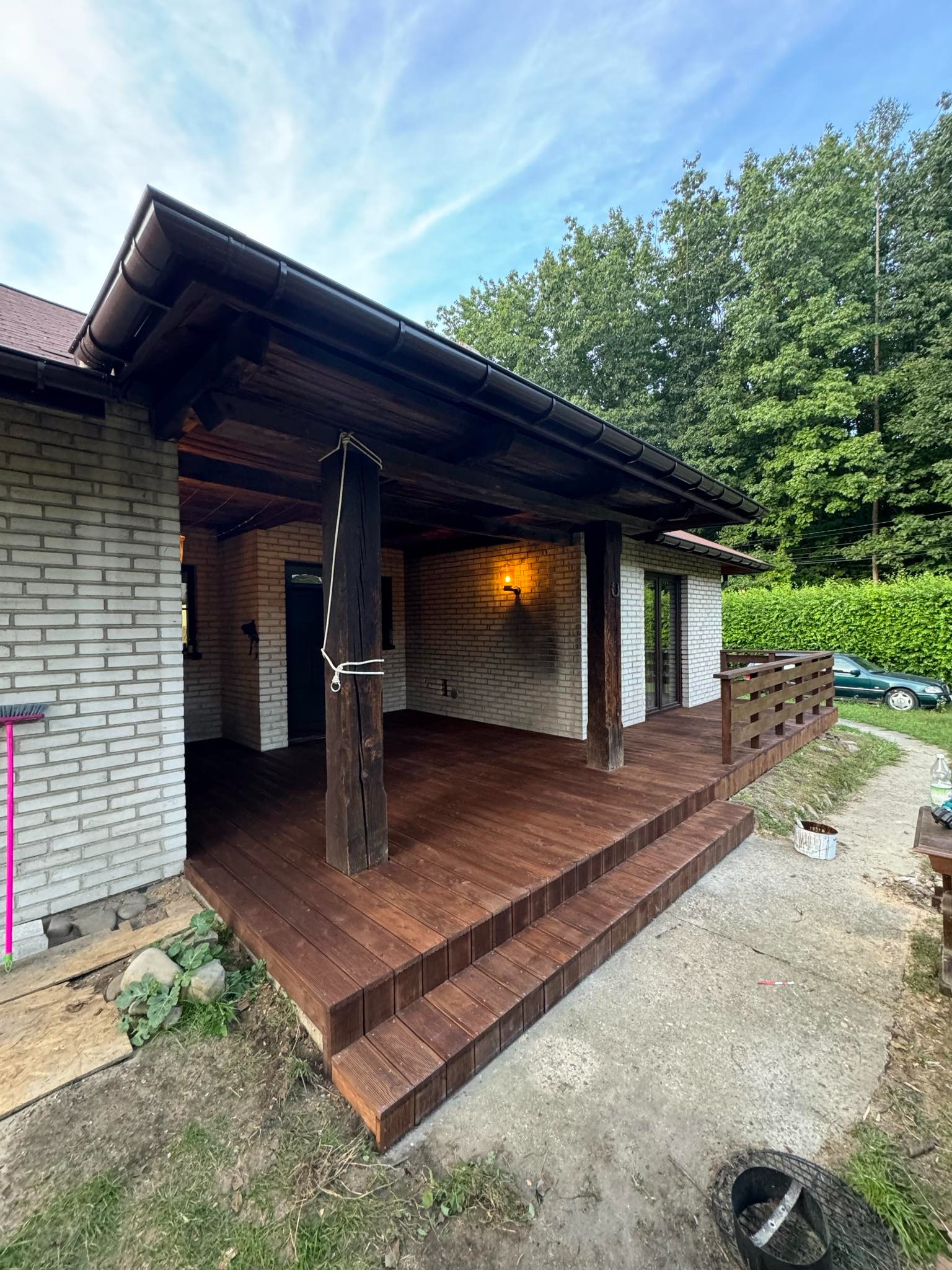 Zadaszony taras z drewnianą podłogą i schodami, wsparty na drewnianych kolumnach, z widoczną ścianą budynku z białej cegły i zielonym tłem drzew. Na pierwszym planie fragment betonowego podłoża.