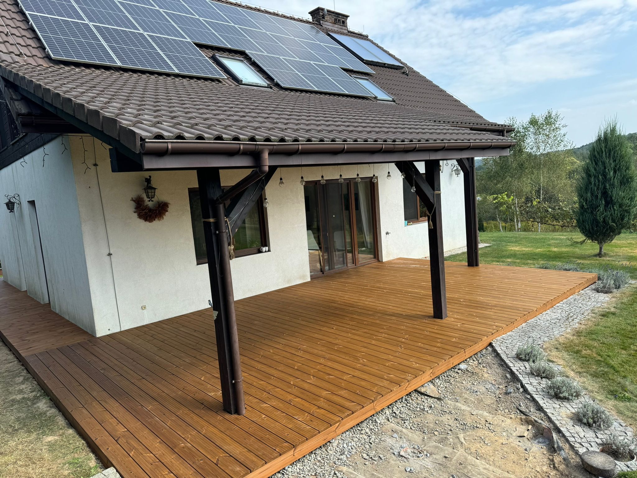 Nowoczesny dom z brązowym dachem i panelami słonecznymi, z drewnianym tarasem zadaszonym belkami, widok na ogród i wzgórza.