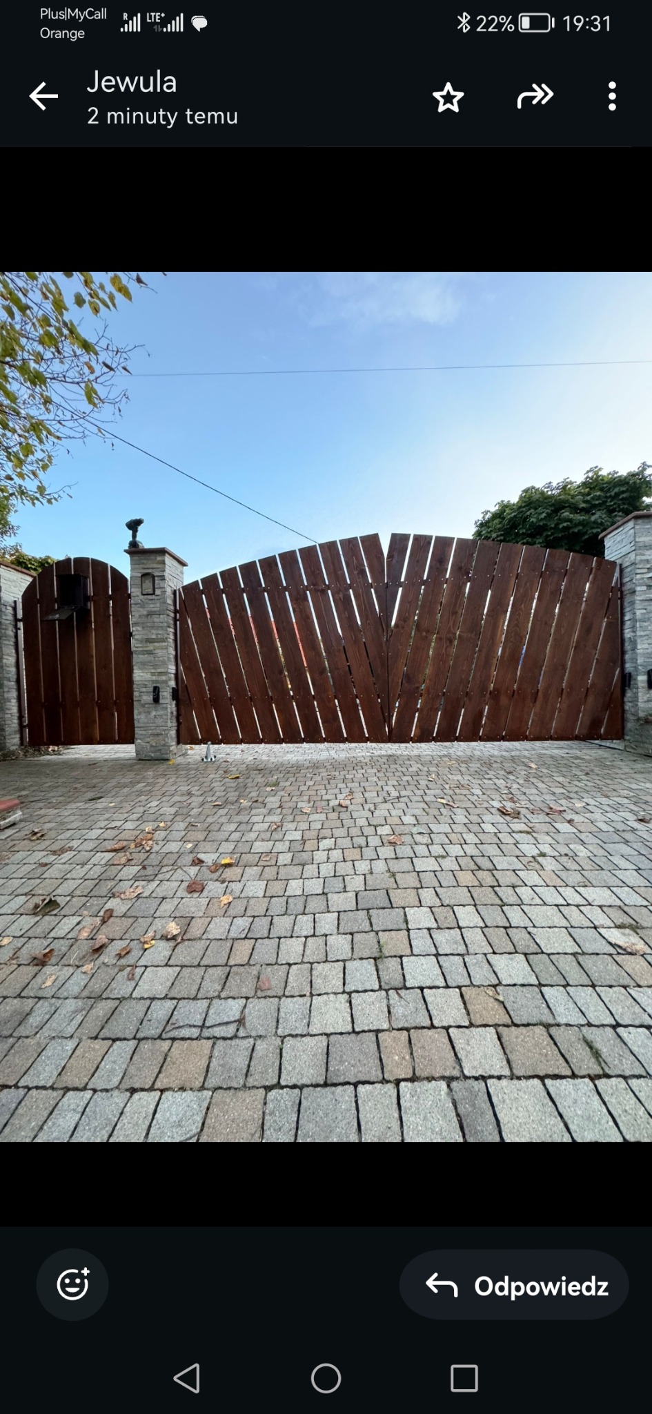 Dwuskrzydłowa, drewniana brama wjazdowa z ukośnymi deskami, osadzona na kamiennych słupkach, z furtką po lewej stronie i brukowanym podjazdem z liśćmi.