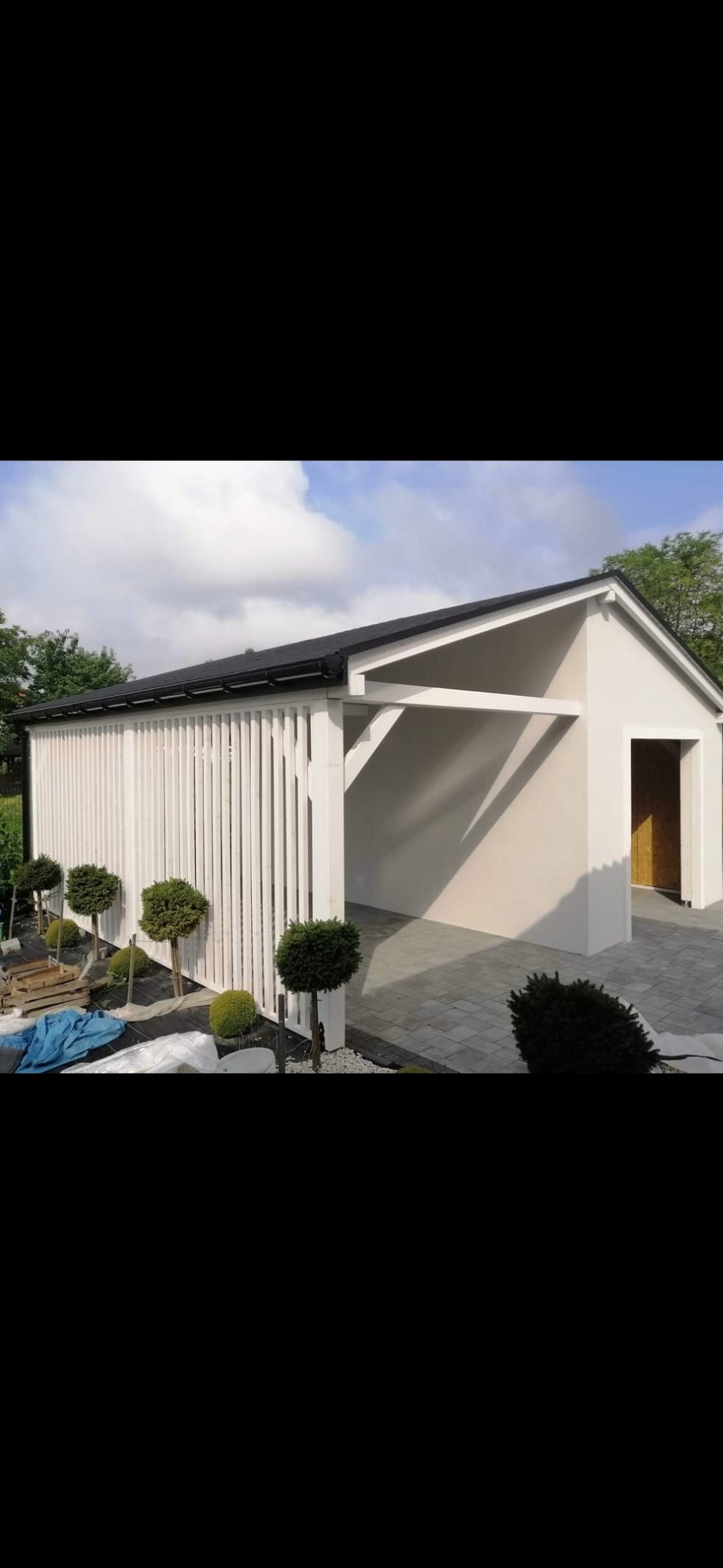 Nowoczesna wiata garażowa z ażurową ścianą z pionowych, białych desek, wykończona kostką brukową i ozdobnymi kulistymi krzewami.