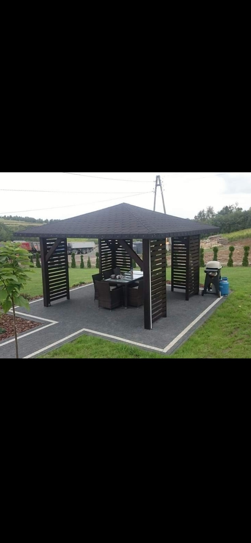 Ciemna, kwadratowa altana z drewnianymi panelami i stołem z krzesłami w środku, ustawiona na szarej kostce brukowej, obok grilla gazowego i trawnika.