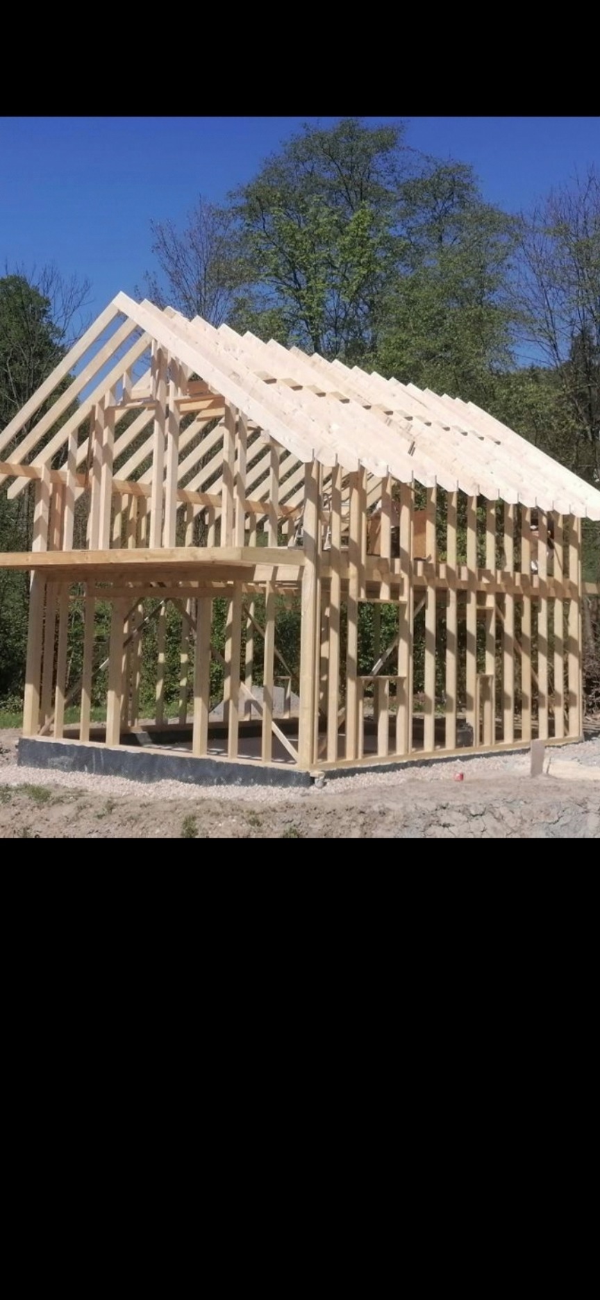 Wznoszenie konstrukcji domu z drewna w Zielonkach, widoczny szkielet budynku z belek i krokwi, na tle drzew i błękitnego nieba.