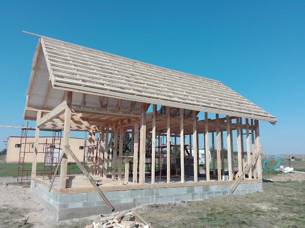 Surowa konstrukcja drewniana budynku z widocznym szkieletem dachu i ścian, osadzona na fundamencie z bloczków betonowych, na tle błękitnego nieba i zielonego krajobrazu.