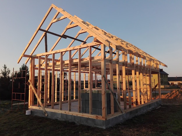 Konstrukcja szkieletowa budynku w trakcie budowy, widoczne drewniane elementy nośne i fundament z bloczków betonowych. W tle budynek i drzewa.