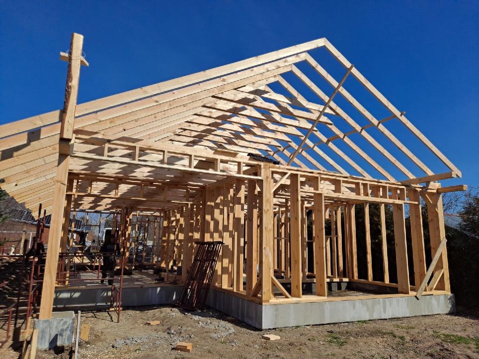 Surowa konstrukcja domu szkieletowego w trakcie budowy, widoczne drewniane ramy ścian i dachu na betonowej podstawie, błękitne niebo w tle, wczesny etap prac.