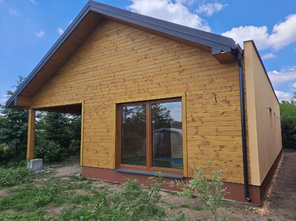 Nowoczesny dom szkieletowy z drewnianą elewacją i zadaszonym tarasem, widok z zewnątrz w słoneczny dzień. Część budynku otynkowana na beżowo.