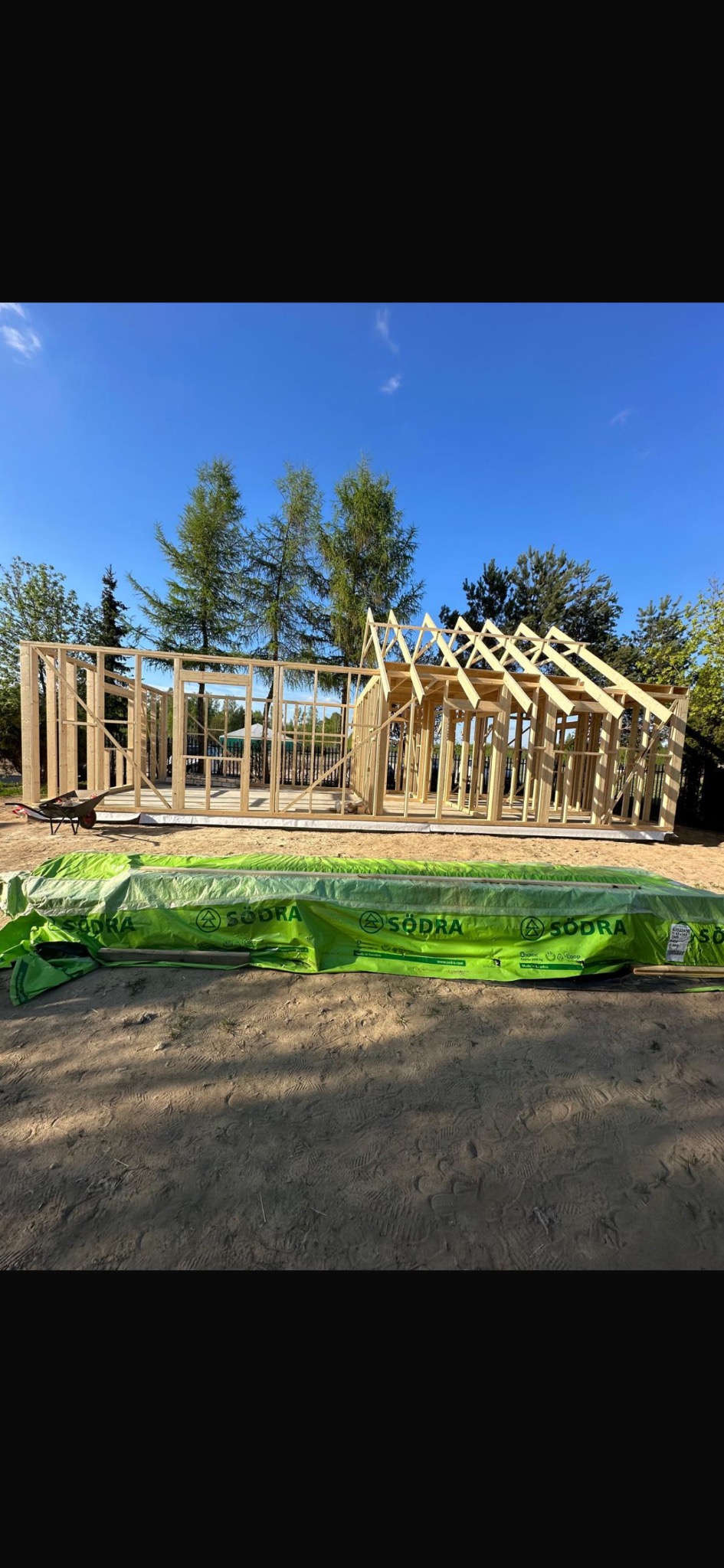 Wznoszenie szkieletu domu z drewna na fundamencie, widoczne elementy konstrukcyjne i materiały budowlane zabezpieczone folią. Ujęcie w słoneczny dzień.