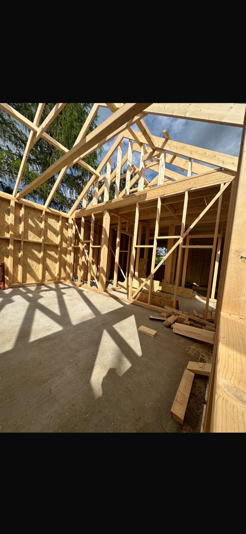 Wnętrze domu w trakcie budowy w technologii szkieletowej. Widoczne drewniane elementy konstrukcyjne ścian i dachu, surowe podłoże i ostre cienie wpadające przez otwory.