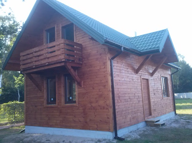 Zgrabny, parterowy dom szkieletowy z drewnianą elewacją w ciepłym odcieniu brązu. Widoczny balkon z drewnianą balustradą i zielony dach z rynnami. Otoczenie: trawnik i drzewa.
