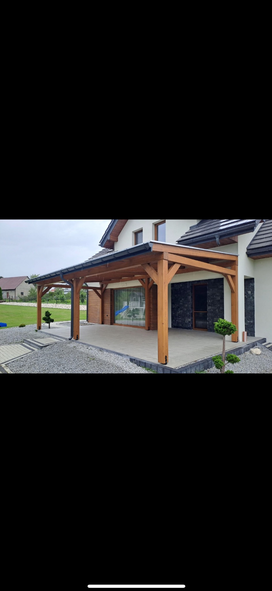 Drewniana pergola zadaszająca taras przy domu jednorodzinnym. Widoczne elementy konstrukcyjne, wykończenie tarasu płytami i otoczenie z kamienia ozdobnego.