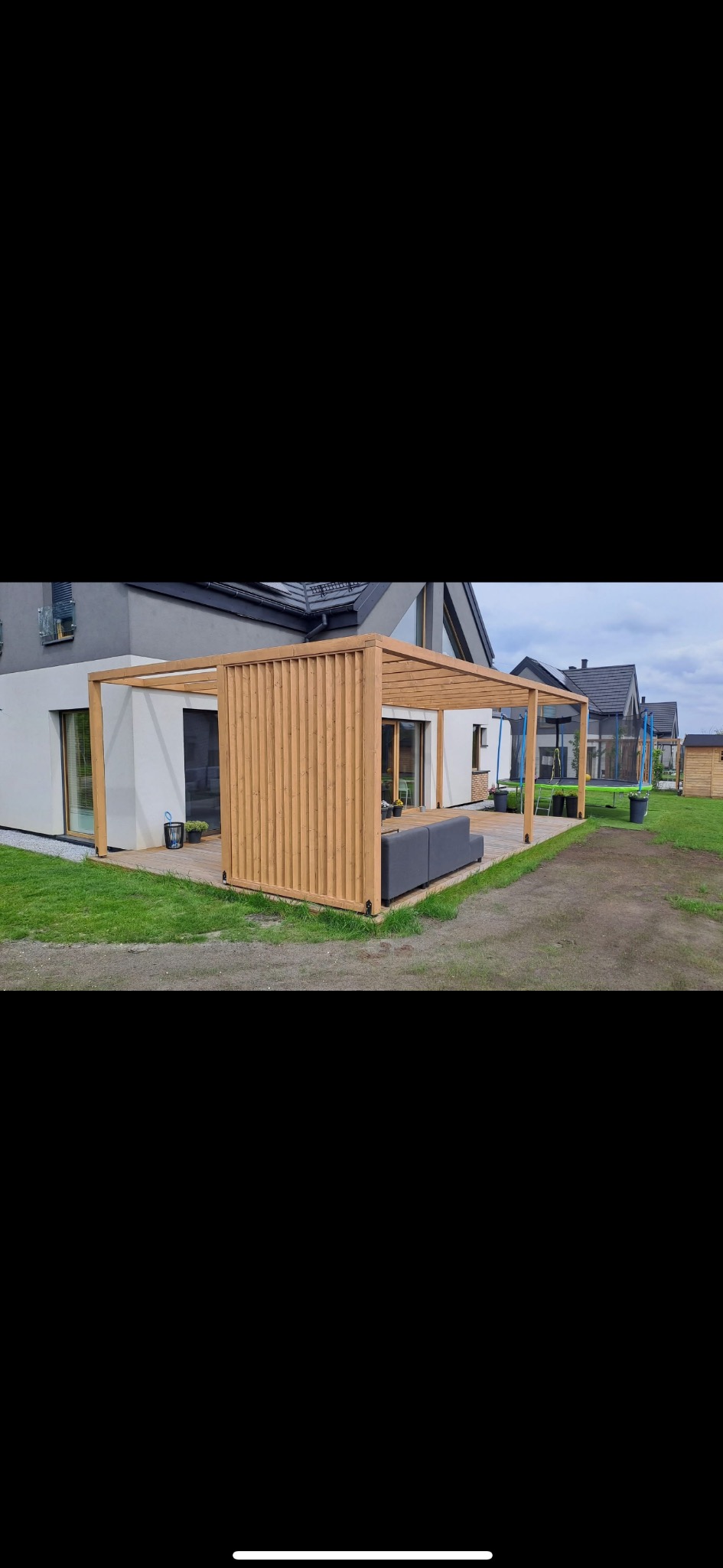 Nowoczesna pergola z jasnego drewna, przylegająca do domu z dużymi oknami. Na tarasie szara sofa, w tle trawnik i inne domy. Widok z boku, w dzień.