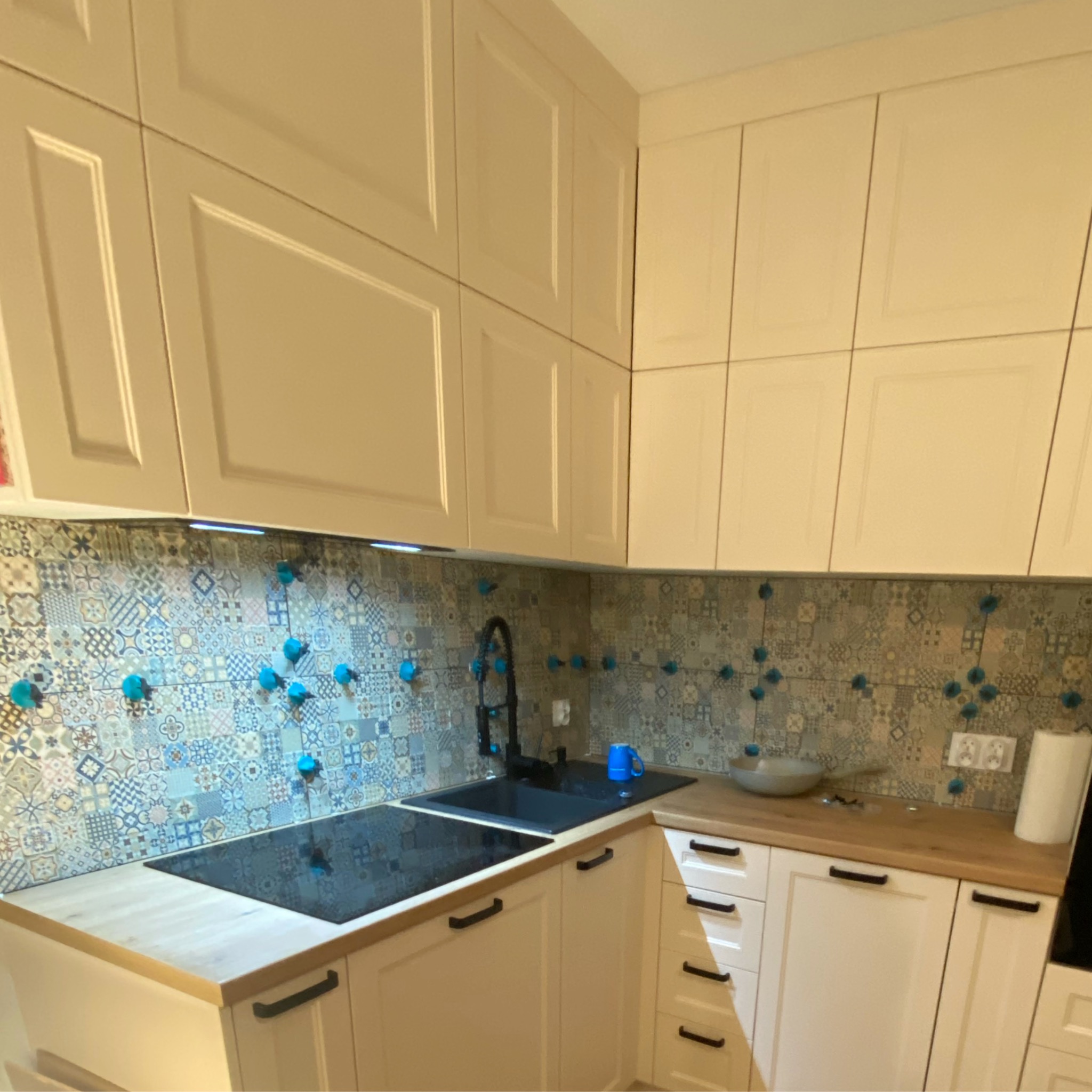 Kuchnia w trakcie montażu płytek ceramicznych na ścianie nad blatem, widoczne niebieskie przyssawki mocujące płytki, jasne szafki kuchenne z czarnymi uchwytami.