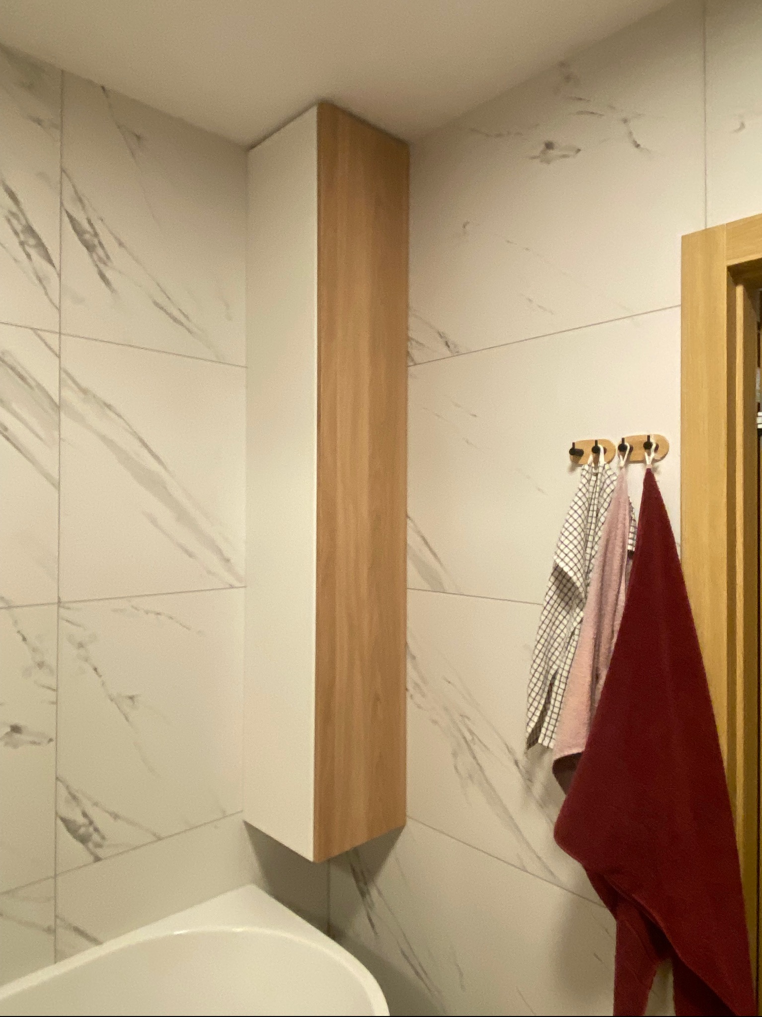 Nowoczesna łazienka z białymi płytkami imitującymi marmur, wisząca szafka z frontem w kolorze drewna, oraz wieszak z trzema ręcznikami w odcieniach bieli, różu i bordo.