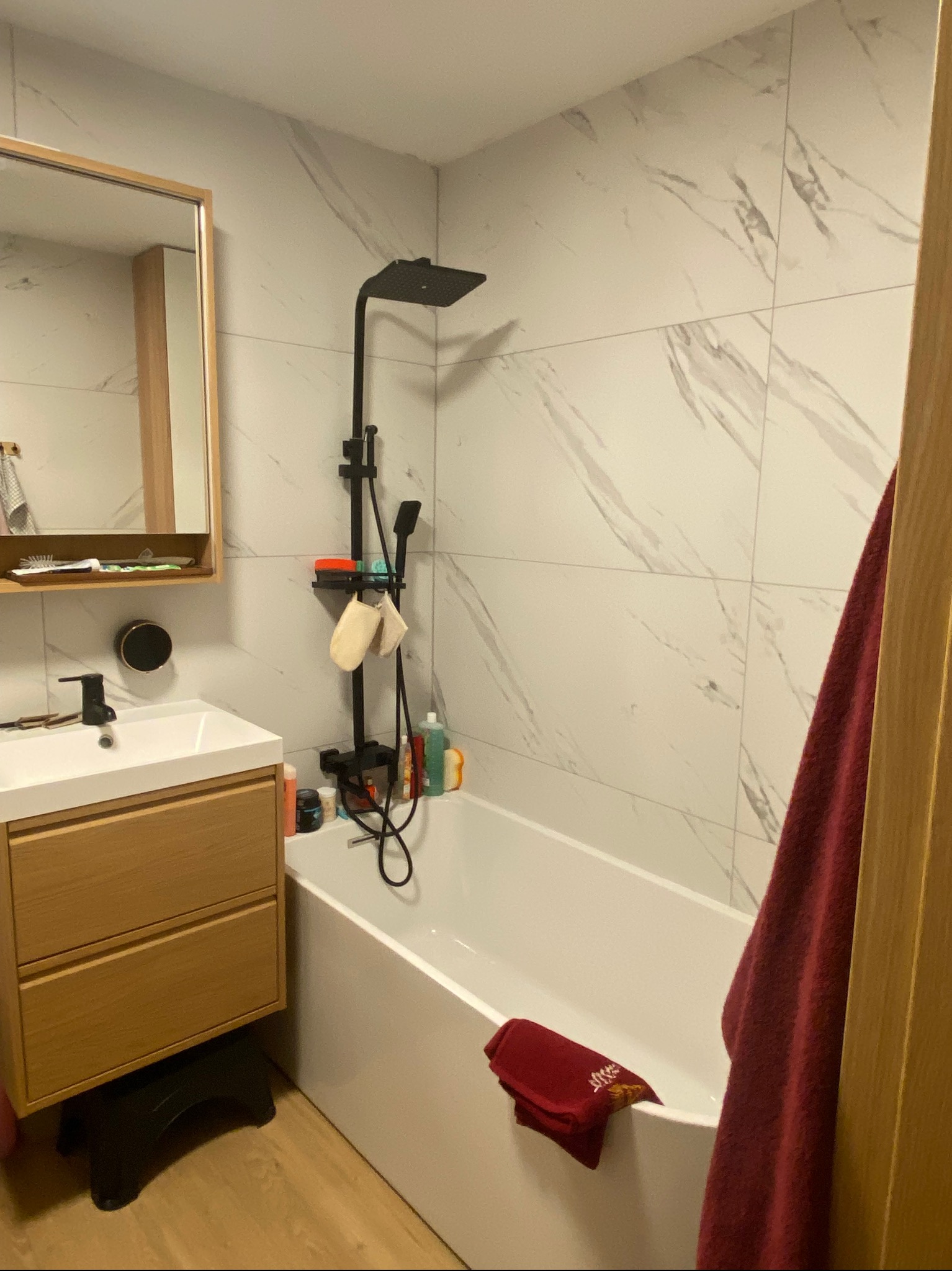 Nowoczesna łazienka z wanną, czarną armaturą i płytkami imitującymi marmur, widok na umywalkę z szafką podwieszaną i lustro.