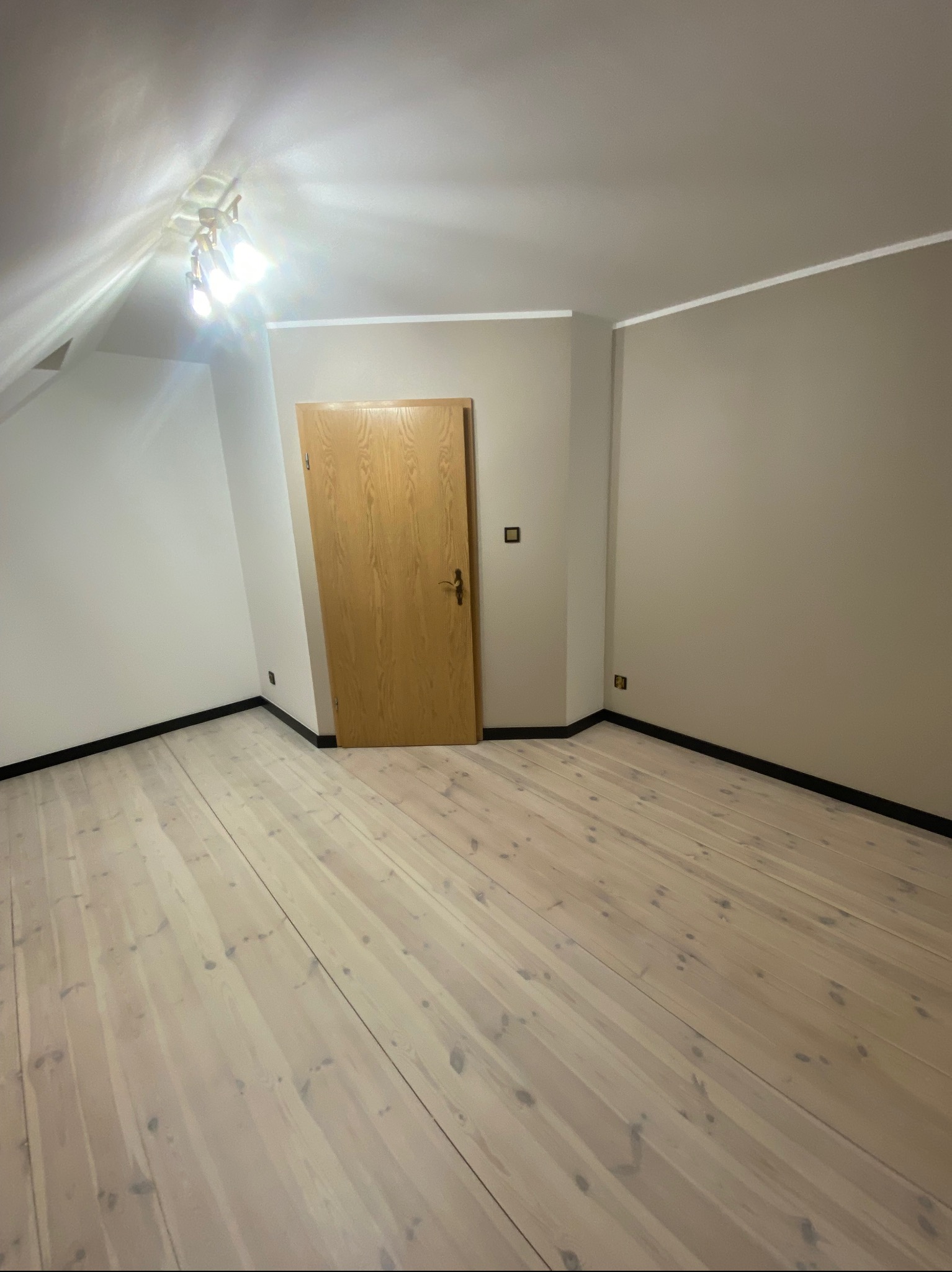 Poddasze po remoncie: świeżo pomalowane ściany w dwóch odcieniach beżu, jasna drewniana podłoga z czarnymi listwami przypodłogowymi i drewniane drzwi.