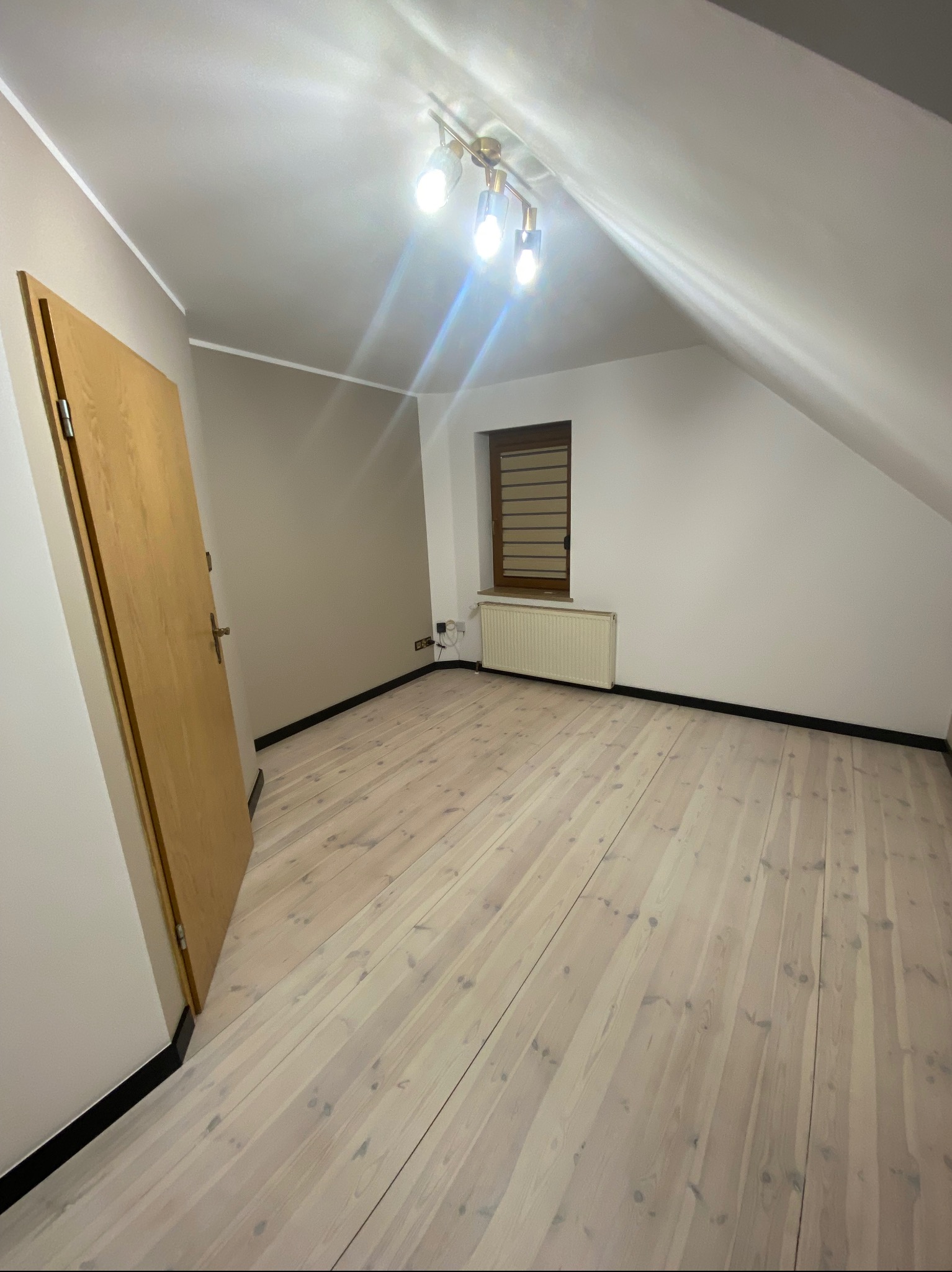 Poddasze po remoncie: jasne panele podłogowe, białe ściany, okno z żaluzjami, grzejnik i złota lampa sufitowa.