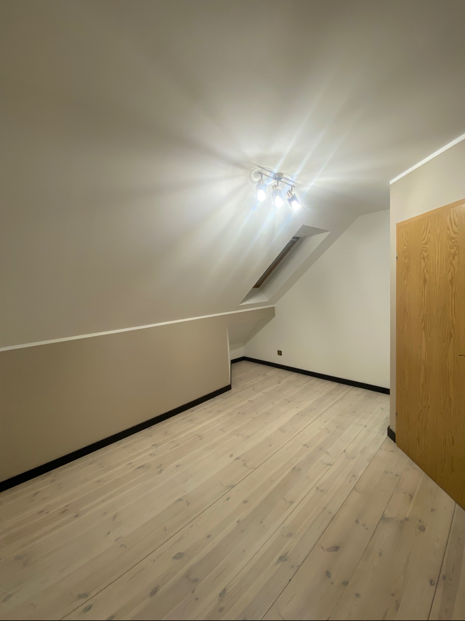 Poddasze po remoncie z jasną drewnianą podłogą, pomalowanymi ścianami w odcieniach beżu i bieli, oświetlone lampą sufitową z trzema reflektorami, widoczne okno dachowe i drewniane drzwi.