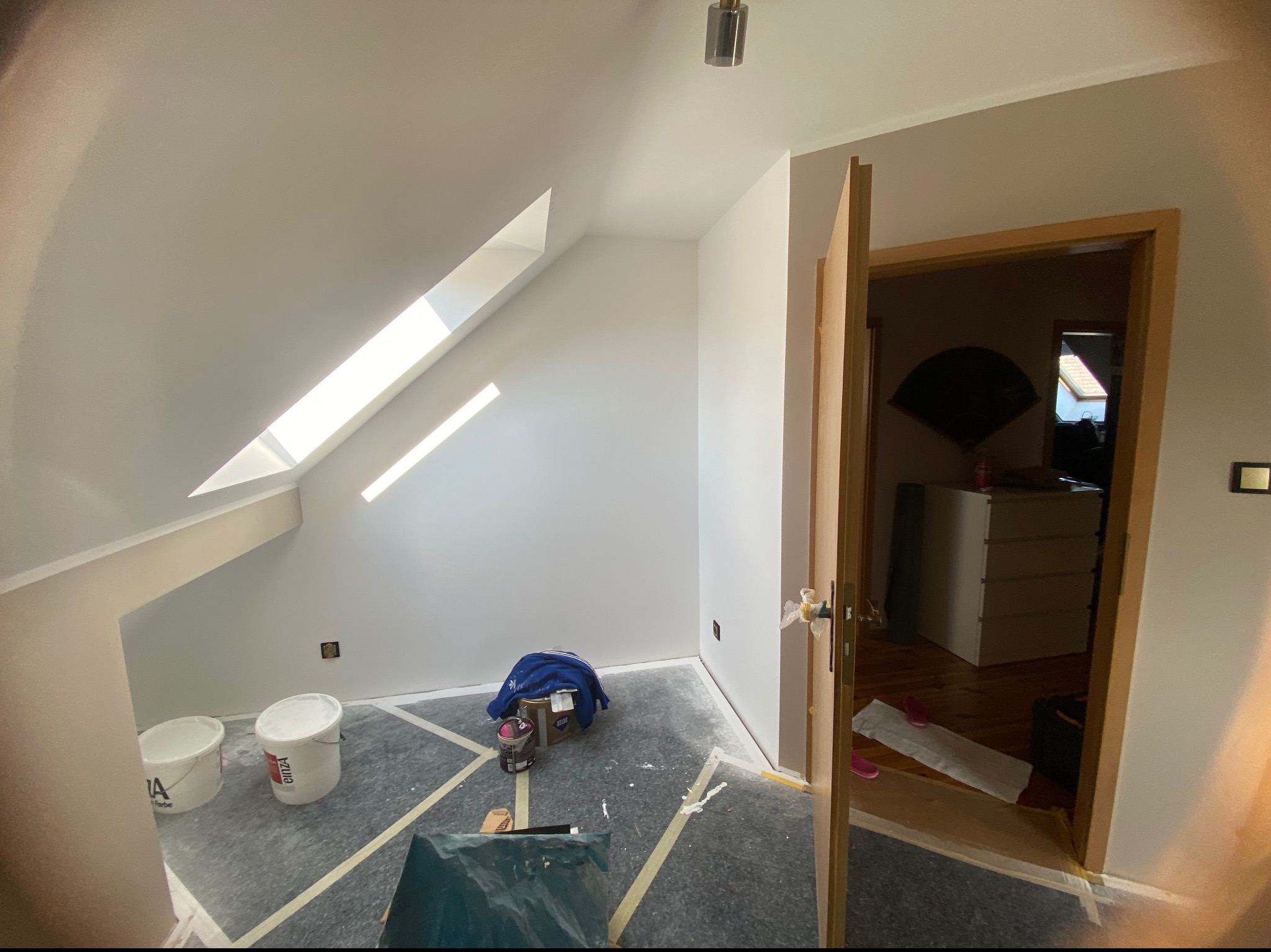 Pokój na poddaszu w trakcie odświeżania: widoczne białe ściany po malowaniu, zabezpieczona folią podłoga, puszki z farbą i narzędzia malarskie. Światło dzienne wpada przez okno dachowe.