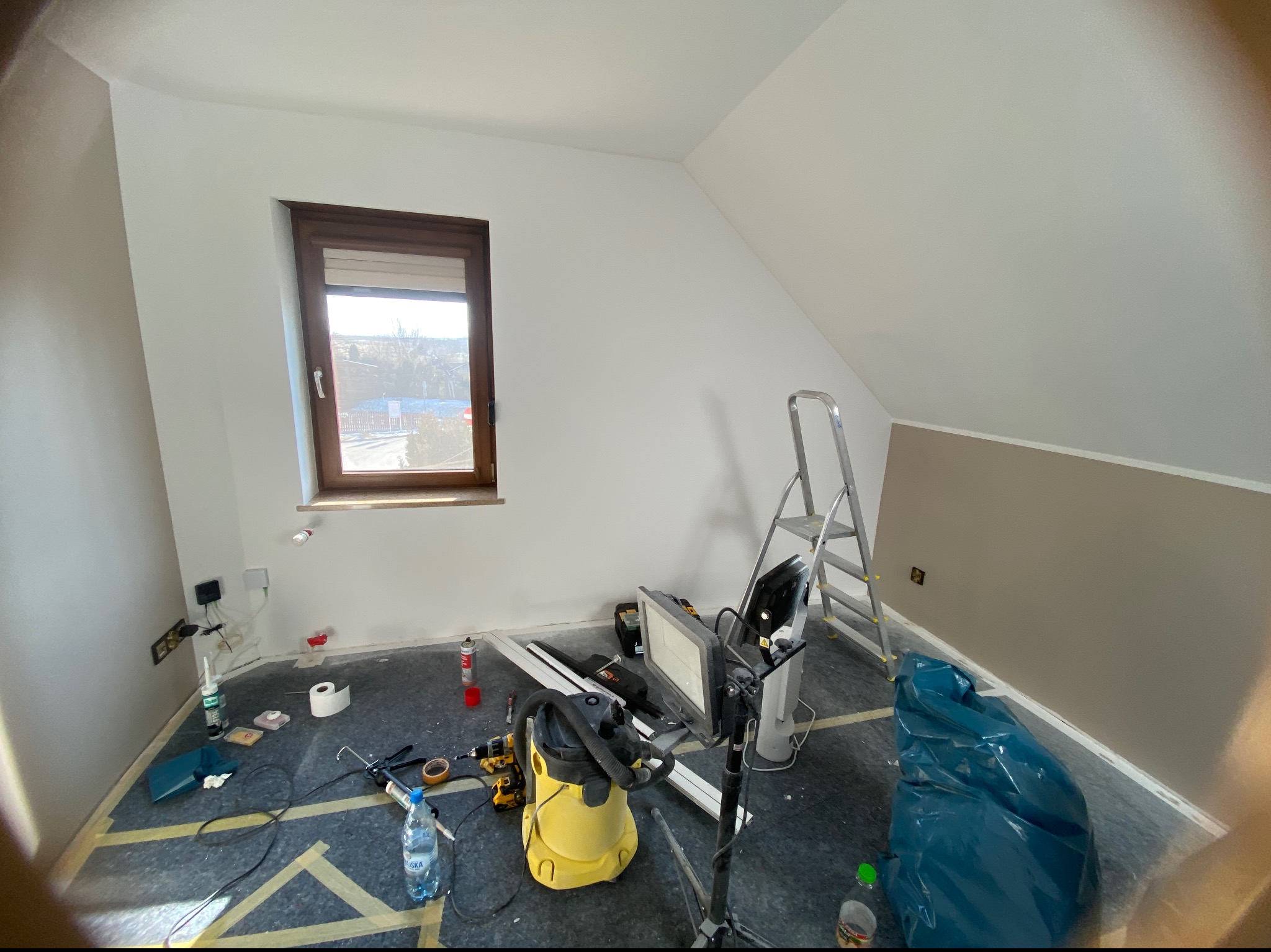 Pokój na poddaszu w trakcie prac wykończeniowych, widoczne narzędzia, drabina, żółty odkurzacz, okno i przygotowane ściany do malowania.