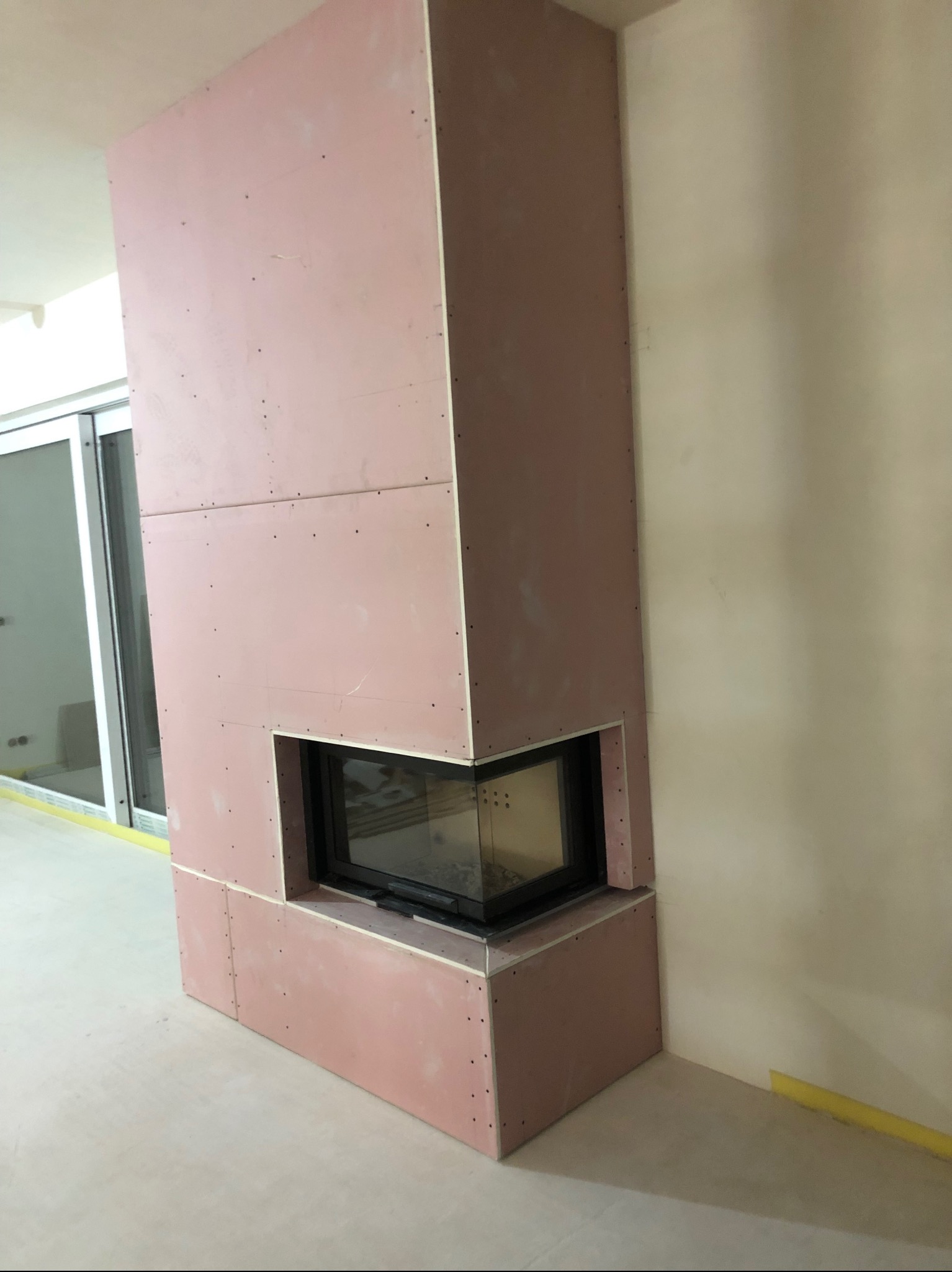 Obudowa kominka z płyty karton gips, na etapie szpachlowania, widoczne łączenia płyt, narożnik z czarnym wkładem kominkowym, zabezpieczenie folią malarską przy podłodze.