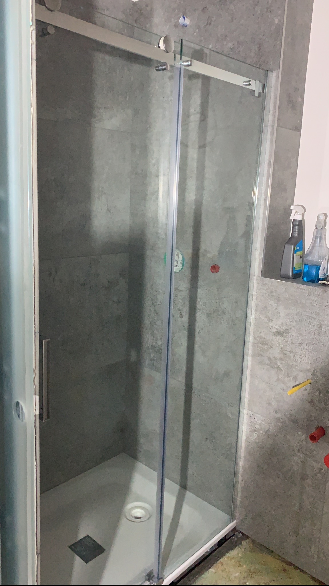 Szklana kabina prysznicowa z przesuwnymi drzwiami na tle szarych płytek, widoczne butelki ze środkami czyszczącymi i fragment instalacji wodnej.