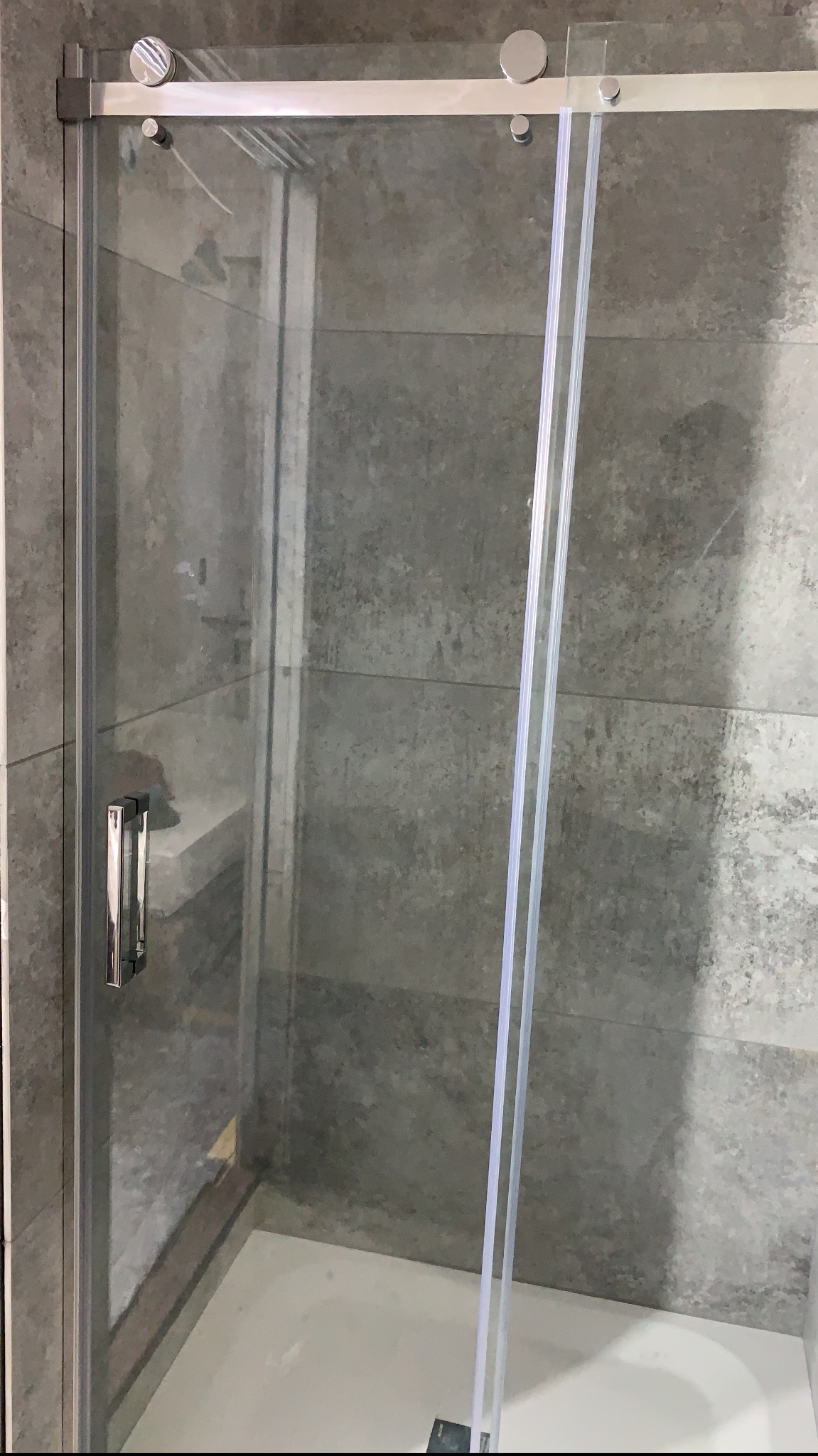 Szklana kabina prysznicowa z przesuwnymi drzwiami, widoczna chromowana listwa i uchwyt, szare płytki na ścianach.