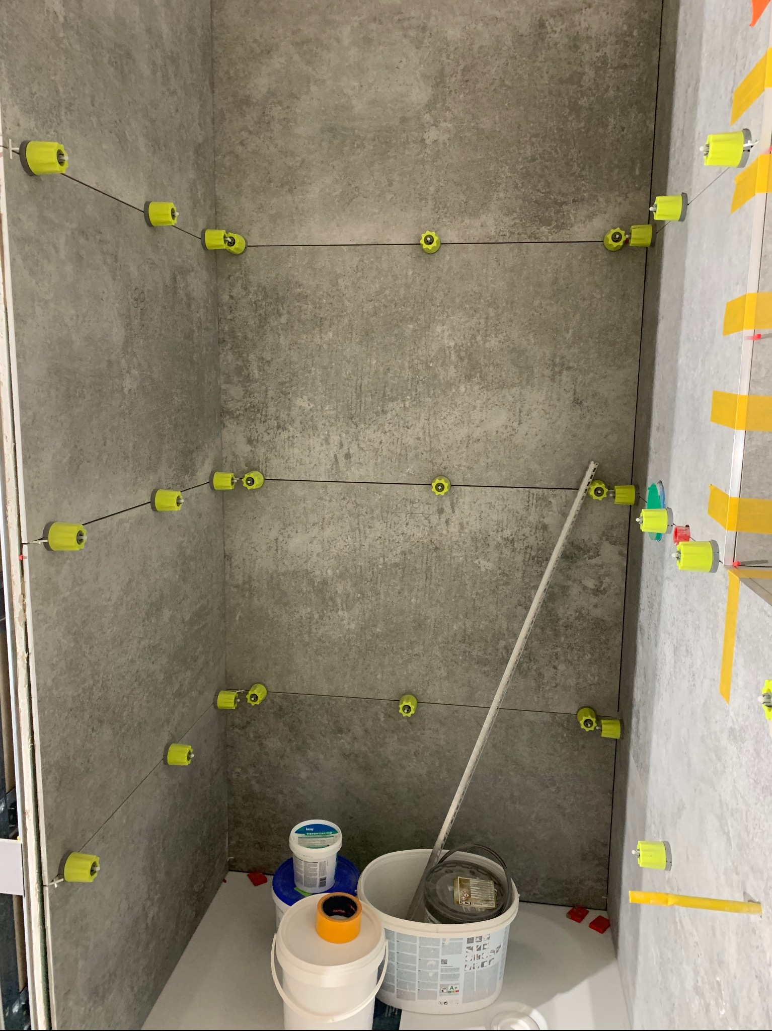 Świeżo ułożone szare płytki ścienne z systemem poziomowania płytek (żółte klipsy i kliny) oraz pojemnikami z klejem w rogu pomieszczenia.