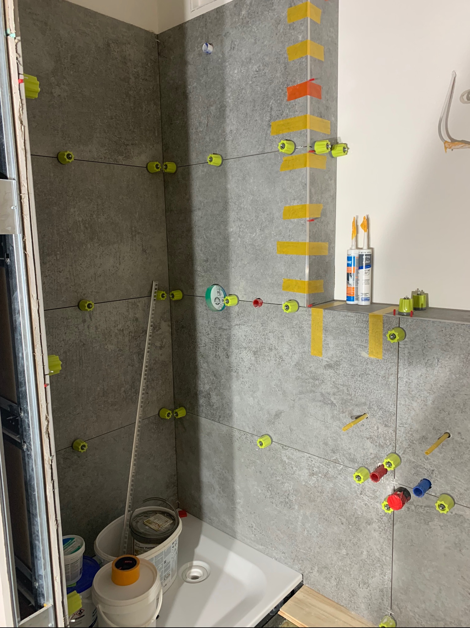 Montaż szarych płytek w kabinie prysznicowej z użyciem systemu poziomowania płytek z żółtymi klipsami i czerwono-niebieskimi klinami, widoczne narzędzia i kleje.