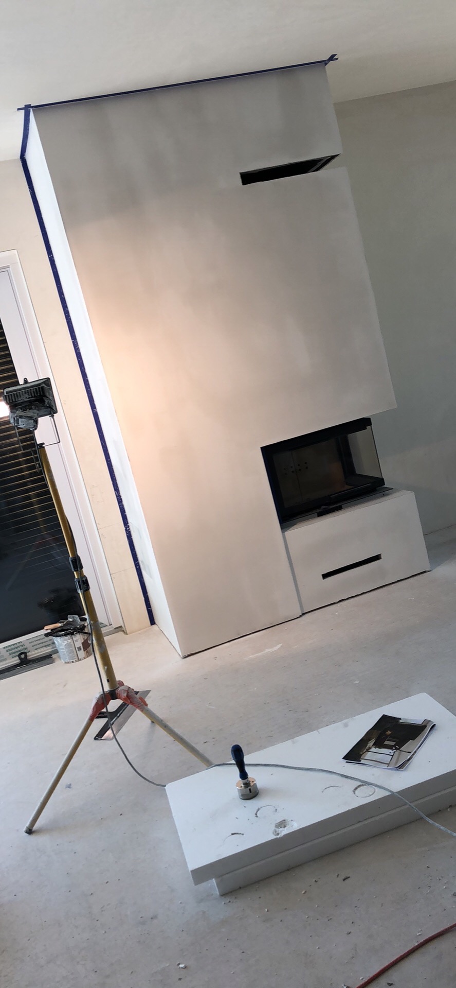 Nowoczesny kominek obudowany płytami, przygotowany do dalszych prac wykończeniowych, zabezpieczony taśmą malarską, oświetlony lampą budowlaną na trójnogu, z widocznym fragmentem okna i podłogą...