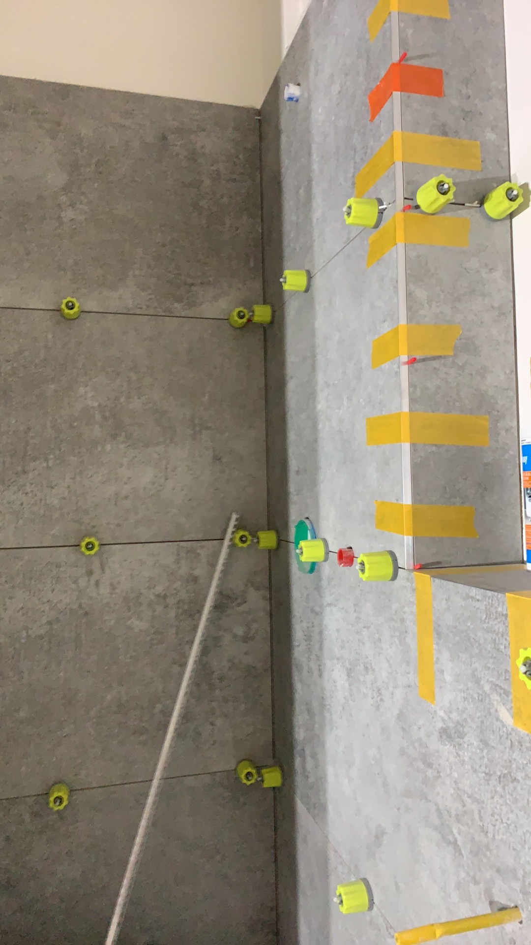 Szare płytki ścienne w trakcie montażu, poziomowanie za pomocą klipsów i klinów, na ścianie widoczne taśmy maskujące.